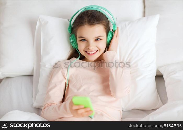 people, children, rest, and technology concept - happy smiling girl lying awake with smartphone and headphones in bed listening to music at home