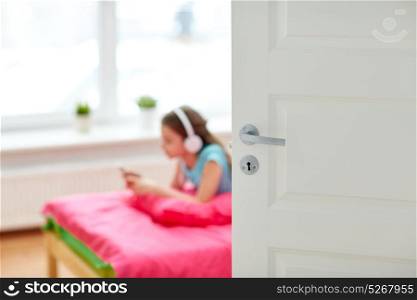 people, children, technology and leisure concept - girl with headphones lying on bed and listening to music at home behind door. girl in headphones listening to music at home