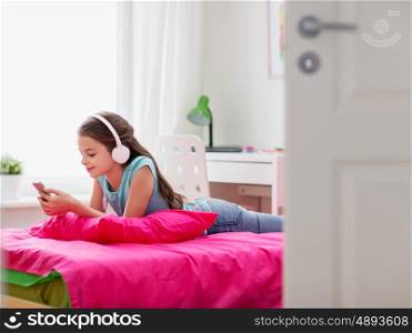 people, children, technology and leisure concept - happy girl with headphones and smartphone lying on bed and listening to music at home
