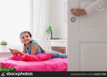 people, children, technology and leisure concept - happy girl with headphones and smartphone lying on bed and listening to music at home, mother opens door in her room
