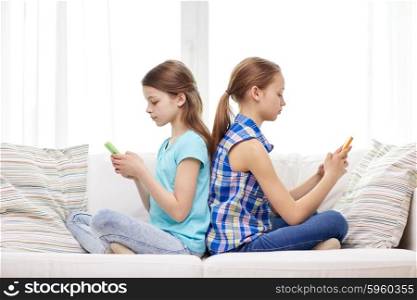 people, children, technology, friends and addiction concept - little girls with smartphones sitting on sofa back to back at home