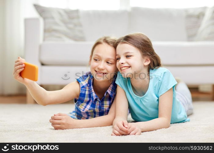 people, children, technology, friends and friendship concept - happy little girls lying on floor and taking selfie with smartphone at home