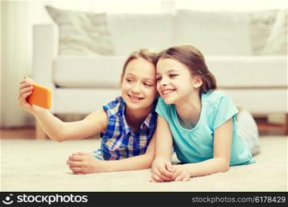 people, children, technology, friends and friendship concept - happy little girls lying on floor and taking selfie with smartphone at home