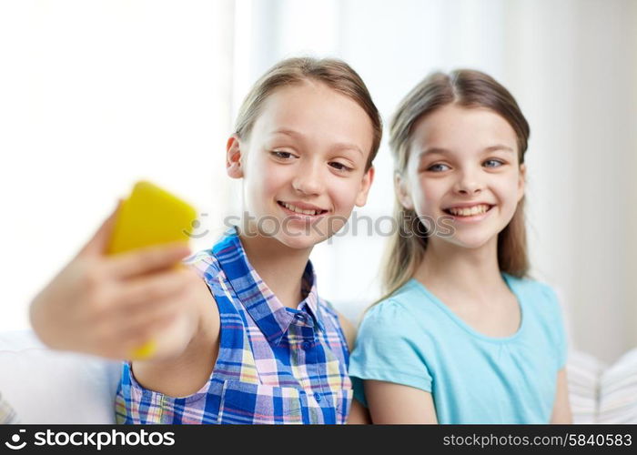 people, children, technology, friends and friendship concept - happy little girls sitting on sofa and taking selfie with smartphone at home