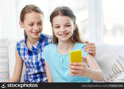 people, children, technology, friends and friendship concept - happy little girls sitting on sofa and taking selfie with smartphone and hugging at home