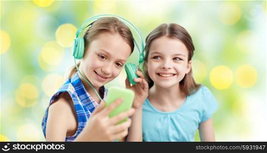 people, children, technology, friends and friendship concept - happy little girls with smartphone and earphones sitting on sofa and listening to music over green lights background
