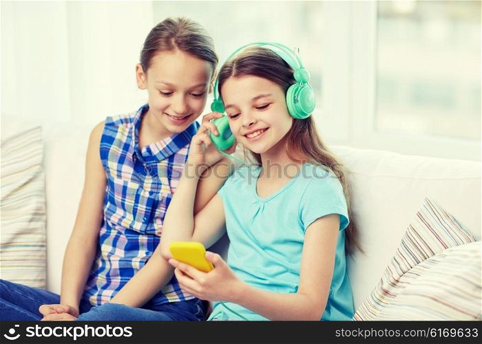 people, children, technology, friends and friendship concept - happy little girls with smartphone and earphones sitting on sofa and listening to music at home