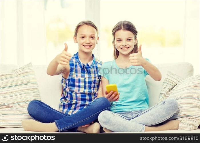 people, children, technology, friends and friendship concept - happy little girls with smartphone sitting on sofa and showing thumbs up at home