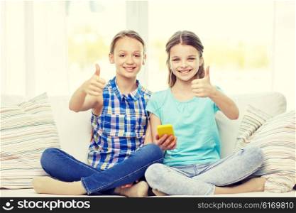 people, children, technology, friends and friendship concept - happy little girls with smartphone sitting on sofa and showing thumbs up at home