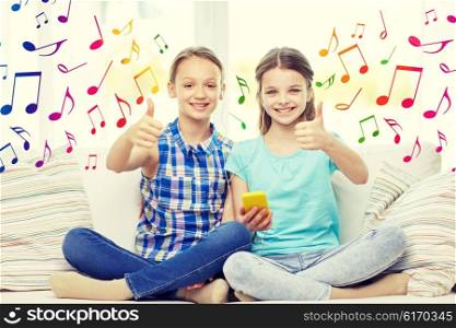 people, children, technology, friends and friendship concept - happy little girls with smartphone and earphones listening to music and showing thumbs up at home over colorful musical notes background