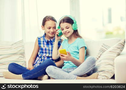 people, children, technology, friends and friendship concept - happy little girls with smartphone and earphones sitting on sofa and listening to music at home