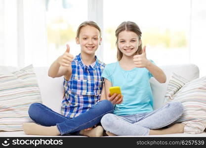 people, children, technology, friends and friendship concept - happy little girls with smartphone sitting on sofa and showing thumbs up at home