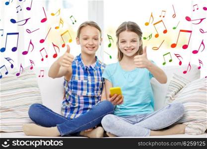 people, children, technology, friends and friendship concept - happy little girls with smartphone and earphones listening to music and showing thumbs up at home over colorful musical notes background