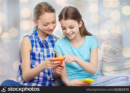 people, children, technology, friends and friendship concept - happy little girls with smartphones sitting on sofa at home over holidays lights background