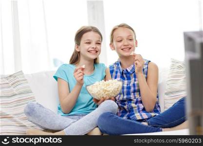 people, children, television, friends and friendship concept - two happy little girls watching comedy movie on tv and eating popcorn at home