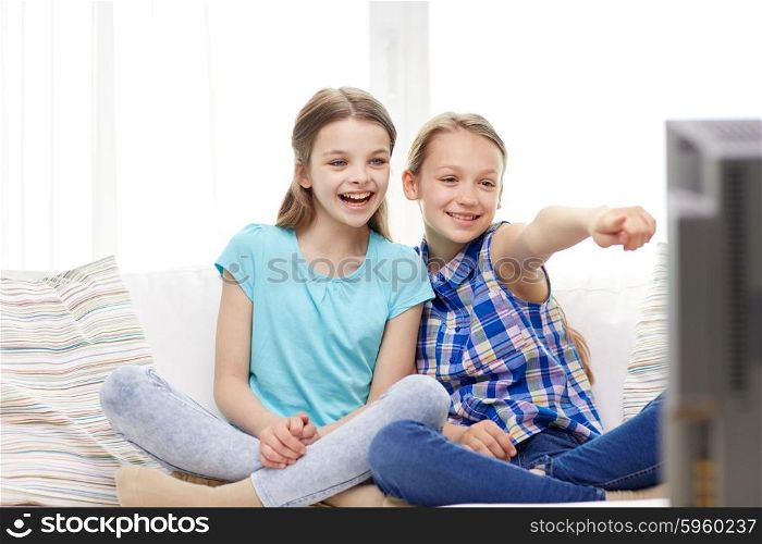 people, children, television, friends and friendship concept - two happy little girls watching tv and pointing finger at home