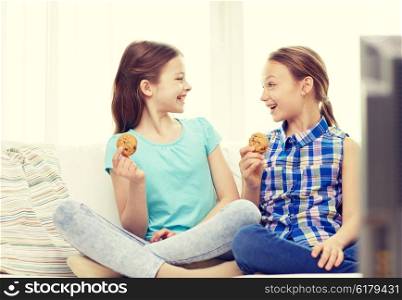 people, children, television, friends and friendship concept - two happy little girls watching tv and eating cookies at home