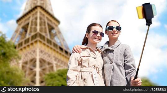 people, children, travel, tourism and technology concept - happy girls taking picture with smartphone on selfie stick over paris eiffel tower background