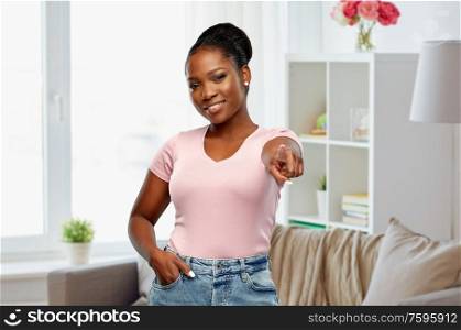 people, choice and gesture concept - happy african american young woman pointing finger to you over home room background. african american woman pointing finger to you