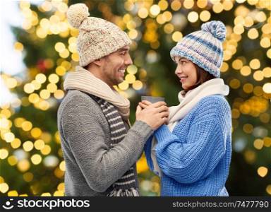 people, christmas and love concept - happy romantic couple in knitted hats and scarves holding one cup over festive lights background. happy couple holding one cup over christmas lights