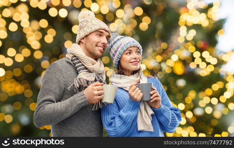 people, christmas and love concept - happy romantic couple in knitted hats and scarves with mugs over festive lights background. happy couple with mugs over christmas lights