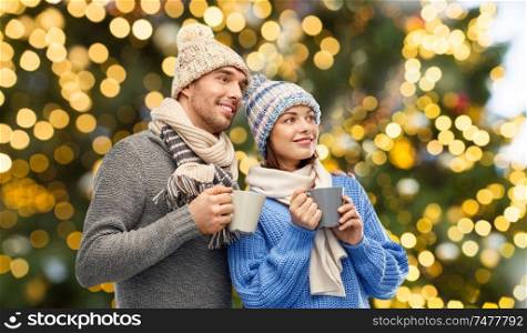 people, christmas and love concept - happy romantic couple in knitted hats and scarves with mugs over festive lights background. happy couple with mugs over christmas lights