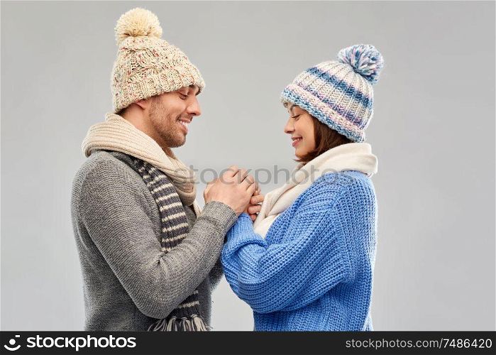 people, christmas and love concept - happy romantic couple in knitted hats and scarves holding hands over grey background. happy couple in winter clothes holding hands