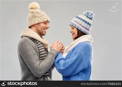 people, christmas and love concept - happy romantic couple in knitted hats and scarves holding hands over grey background. happy couple in winter clothes holding hands