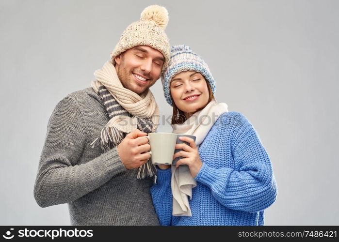 people, christmas and love concept - happy romantic couple in knitted hats and scarves with mugs over grey background. happy couple in winter clothes with mugs