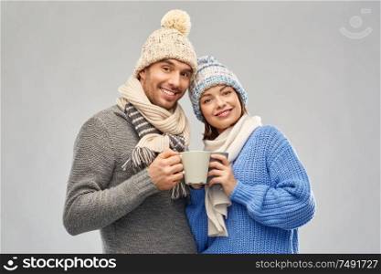 people, christmas and love concept - happy romantic couple in knitted hats and scarves with mugs over grey background. happy couple in winter clothes with mugs