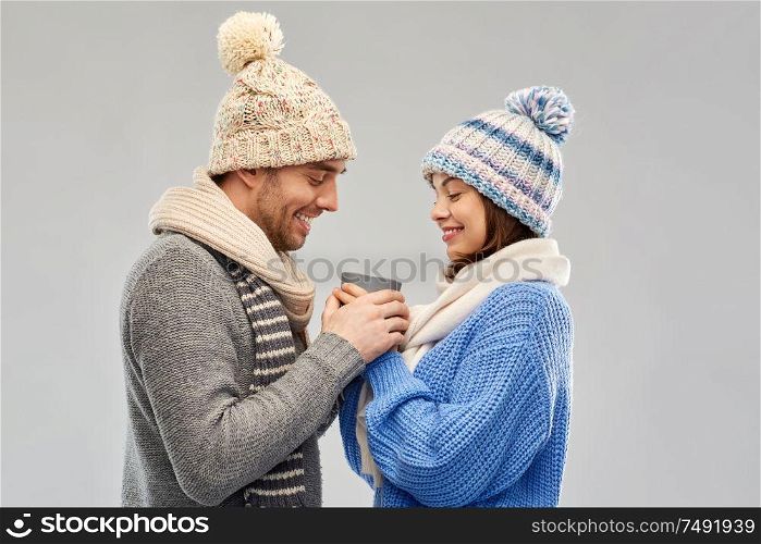 people, christmas and love concept - happy romantic couple in knitted hats and scarves holding one cup over grey background. happy couple in winter clothes holding one cup