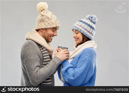 people, christmas and love concept - happy romantic couple in knitted hats and scarves holding one cup over grey background. happy couple in winter clothes holding one cup