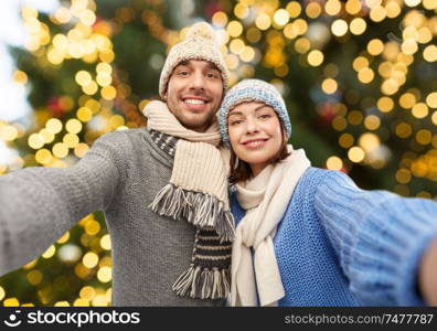 people, christmas and winter clothes concept - happy couple in knitted hats and scarves taking selfie over festive lights background. happy couple taking selfie over christmas lights