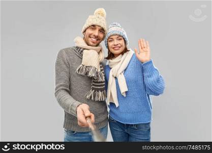 people, christmas and winter clothes concept - happy couple in knitted hats and scarves taking picture by selfie stick over grey background. happy couple in winter clothes taking selfie