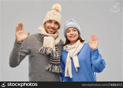 people, christmas and winter clothes concept - happy couple in knitted hats and scarves waving hands over grey background. happy couple in winter clothes waving hands