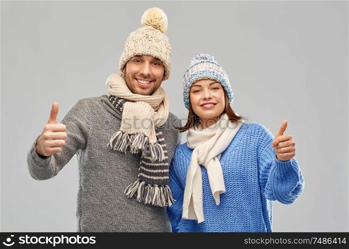 people, christmas and winter clothes concept - happy couple in knitted hats and scarves showing thumbs up over grey background. happy couple in winter clothes showing thumbs up