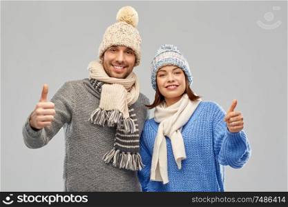 people, christmas and winter clothes concept - happy couple in knitted hats and scarves showing thumbs up over grey background. happy couple in winter clothes showing thumbs up