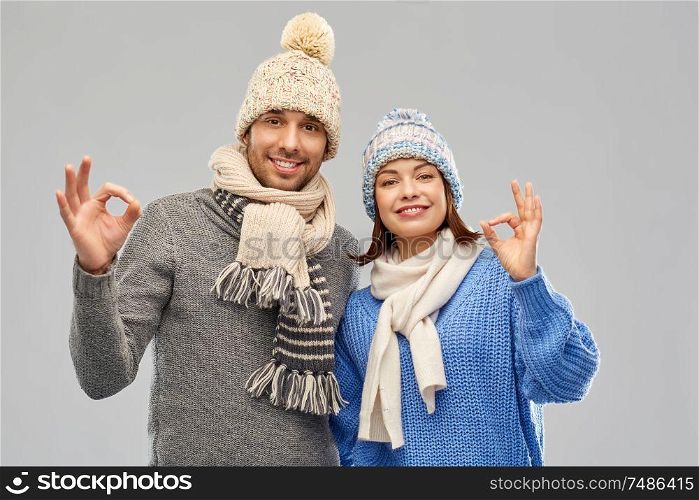 people, christmas and winter clothes concept - happy couple in knitted hats and scarves showing ok hand sign over grey background. happy couple in winter clothes showing ok sign