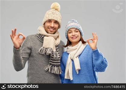 people, christmas and winter clothes concept - happy couple in knitted hats and scarves showing ok hand sign over grey background. happy couple in winter clothes showing ok sign