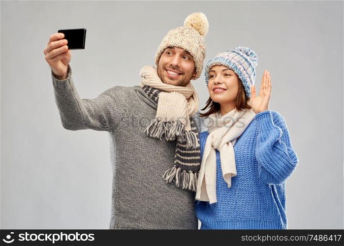 people, christmas and winter clothes concept - happy couple in knitted hats and scarves taking selfie by smartphone over grey background. couple in winter hats taking selfie by smartphone