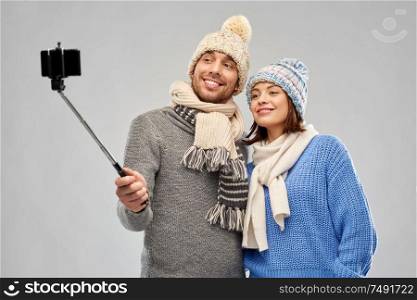 people, christmas and winter clothes concept - happy couple in knitted hats and scarves taking picture by smartphone on selfie stick over grey background. happy couple in winter clothes taking selfie