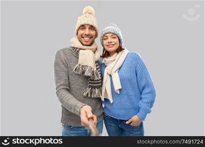 people, christmas and winter clothes concept - happy couple in knitted hats and scarves taking picture by selfie stick over grey background. happy couple in winter clothes taking selfie