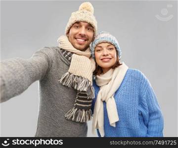 people, christmas and winter clothes concept - happy couple in knitted hats and scarves taking selfie over grey background. happy couple in winter clothes taking selfie