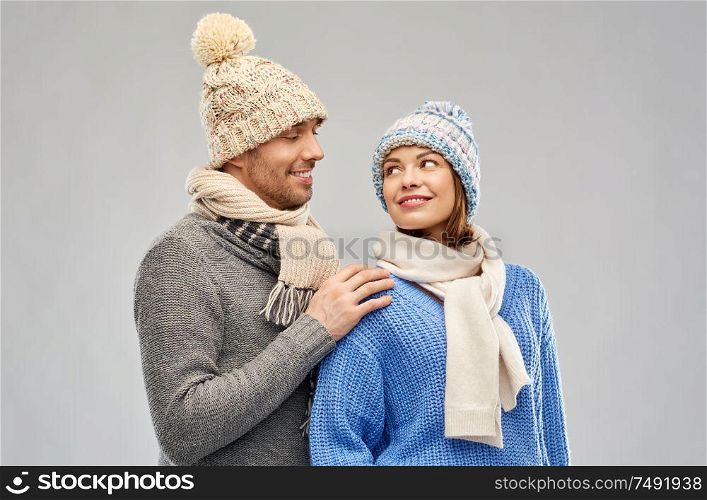 people, christmas and winter clothes concept - happy couple in knitted hats and scarves looking at each other over grey background. happy couple in winter clothes