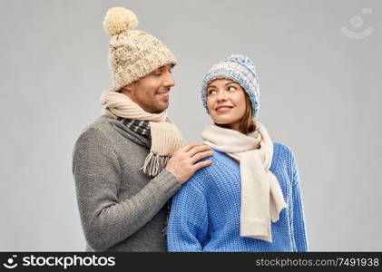 people, christmas and winter clothes concept - happy couple in knitted hats and scarves looking at each other over grey background. happy couple in winter clothes