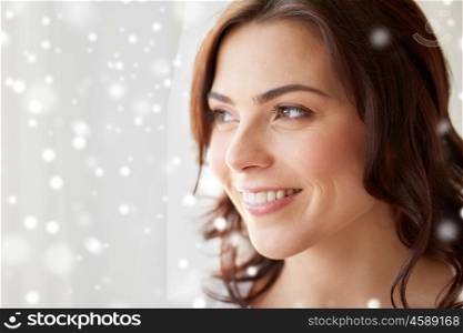 people, christmas and winter concept - close up of happy young woman looking through window at home over snow