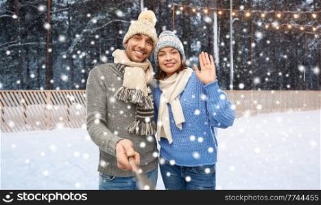 people, christmas and winter holidays concept - happy couple in knitted hats and scarves taking picture by selfie stick over ice rink background. happy couple in winter clothes taking selfie