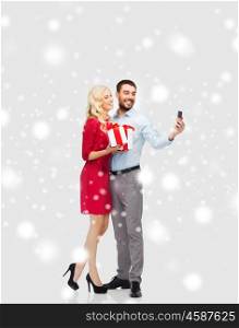 people, christmas, couple, technology and holidays concept - happy young woman and man with birthday presents taking selfie by smartphone over snow background