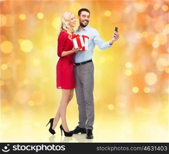 people, christmas, couple, technology and holidays concept - happy young woman and man with birthday presents taking selfie by smartphone over lights background