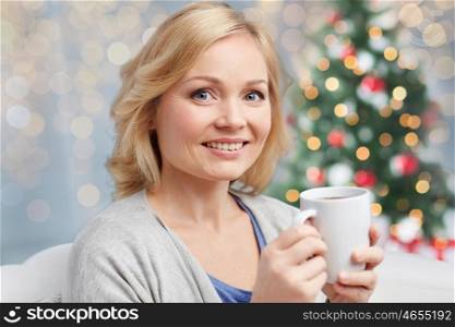 people, christmas, drinks and leisure concept - smiling woman with cup of tea or coffee at home over holidays lights background